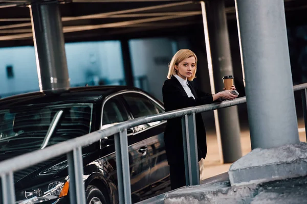 Schönes blondes Mädchen hält Smartphone und Pappbecher in der Hand und steht neben schwarzem Auto — Stockfoto