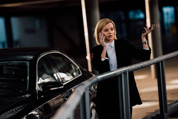 Menina loira atraente falando no smartphone e gestos perto de carro preto — Fotografia de Stock