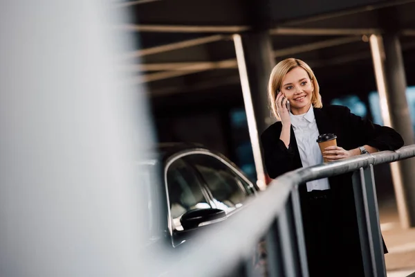 Enfoque selectivo de alegre chica rubia hablando en el teléfono inteligente y sosteniendo taza de papel cerca de coche negro - foto de stock