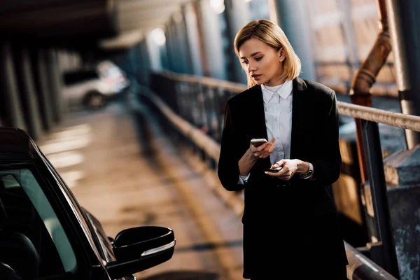 Bela menina loira segurando smartphone e segurando chave perto de carro preto — Fotografia de Stock