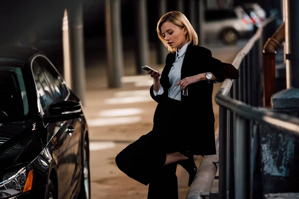 Beautiful blonde girl looking at smartphone and holding key near black car — Stock Photo