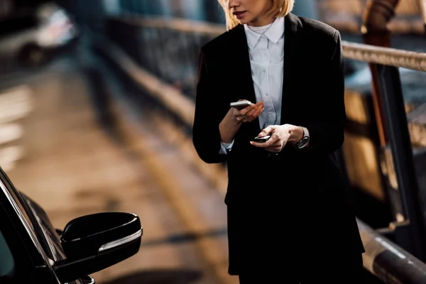 Cropped view of blonde girl holding smartphone and key near black car — Stock Photo