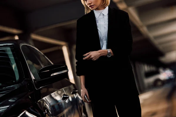 Cropped view of blonde woman holding smartphone and key near black car — Stock Photo