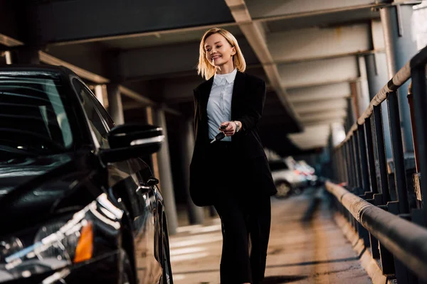 Cheerful attractive blonde woman walking near black automobile in parking — Stock Photo