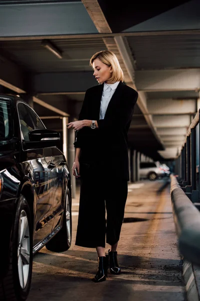 Attractive blonde woman standing near black automobile in parking — Stock Photo