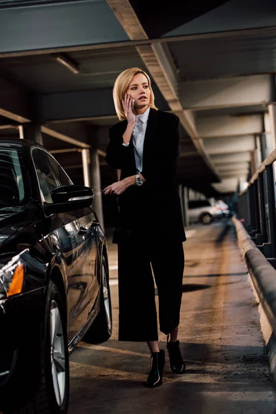 Atractiva mujer rubia de pie cerca de automóvil negro y hablando en el teléfono inteligente - foto de stock