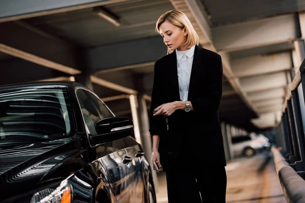 Красивая блондинка держит ключ и стоит рядом с черным автомобилем на парковке — стоковое фото