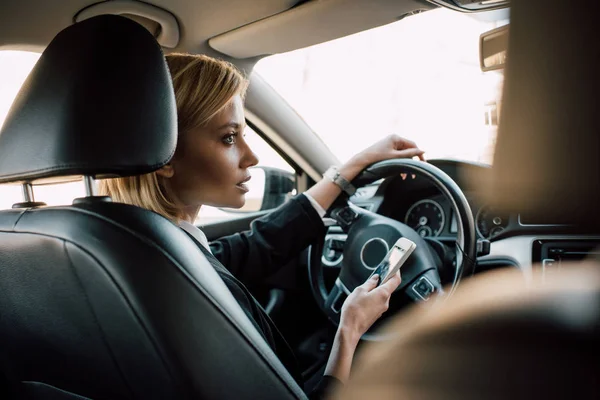 Mulher loira atraente sentado no carro e segurando smartphone — Fotografia de Stock