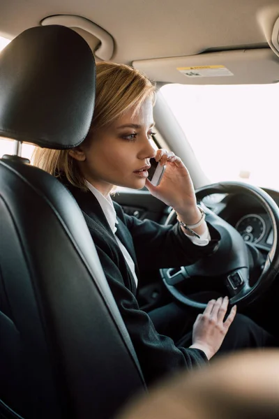 Atractiva rubia joven sentada en el coche y hablando en el teléfono inteligente - foto de stock
