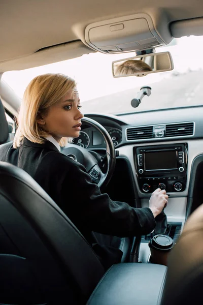 Worried blonde young woman holding gear shift handle while sitting in car — Stock Photo