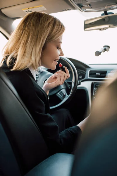 Cheerful blonde girl applying lipstick while sitting in car — Stock Photo