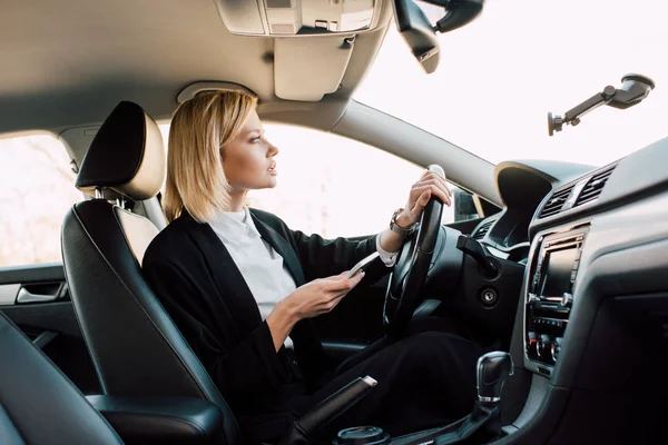 Attraente bionda giovane conducente in possesso di smartphone durante la guida auto — Foto stock