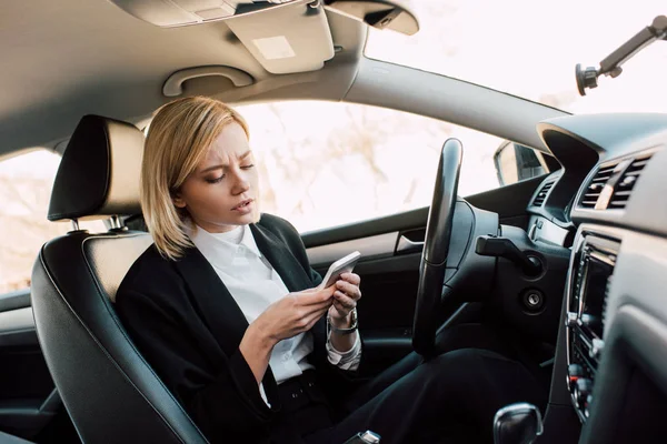 Attractive blonde young woman looking at smartphone while sitting in car — Stock Photo