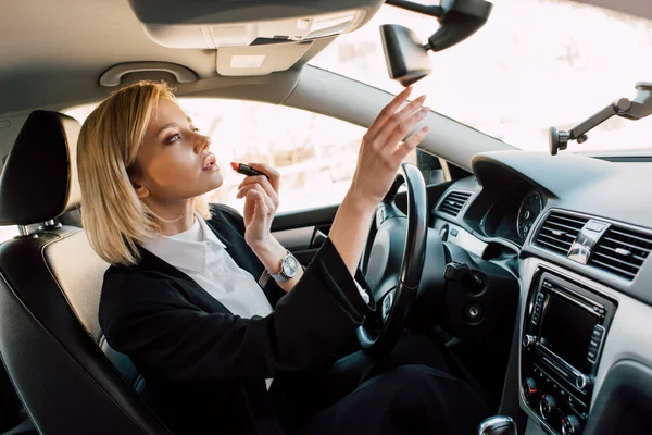 Attractive blonde young woman applying lipstick while sitting in car — Stock Photo