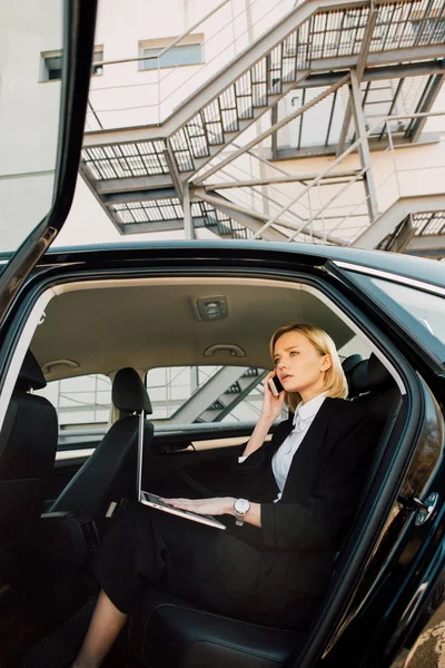 Vista de ángulo bajo de la mujer rubia preocupada hablando en el teléfono inteligente mientras está sentado con el ordenador portátil en el coche - foto de stock