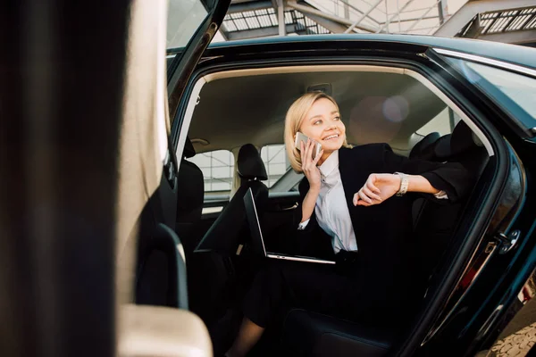 Low angle view of happy woman talking on smartphone while sitting with laptop in car — Stock Photo