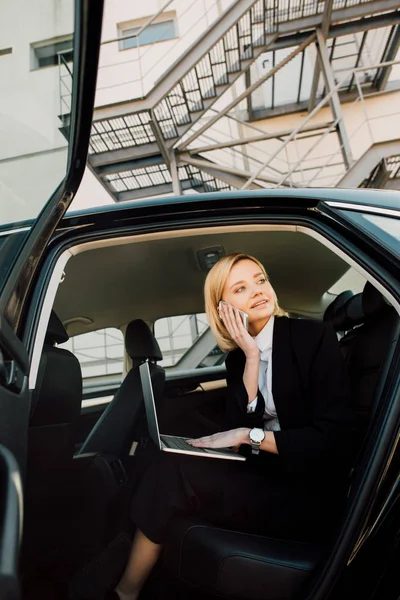 Low angle view of smiling woman talking on smartphone while sitting with laptop in car — Stock Photo