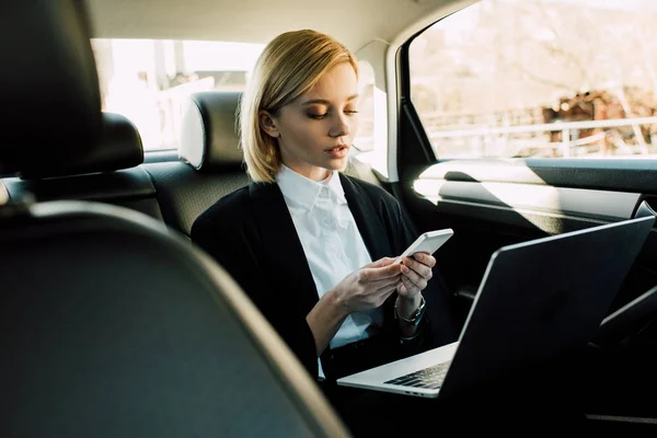 Attraktive blonde junge Frau schaut aufs Smartphone, während sie mit Laptop im Auto sitzt — Stockfoto