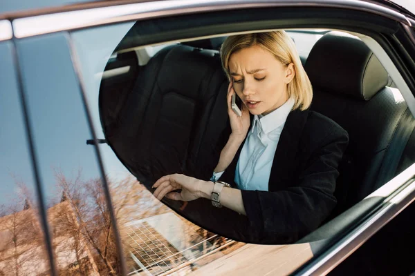 Upset blonde woman talking on smartphone and looking at watch in car — Stock Photo