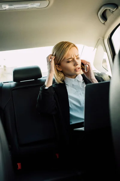 Confused blonde woman talking on smartphone near laptop in car — Stock Photo