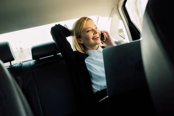 Happy blonde woman talking on smartphone and smiling near laptop in car — Stock Photo