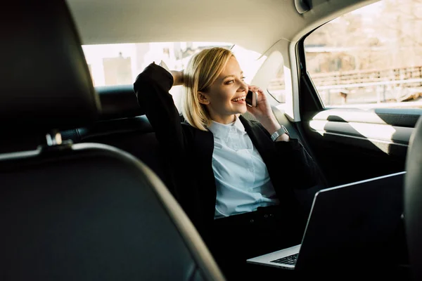 Smiling blonde woman talking on smartphone near laptop in car — Stock Photo