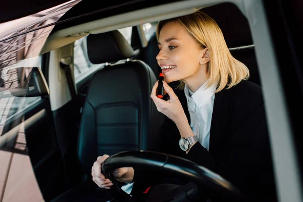 Fröhliche blonde junge Frau trägt Lippenstift im Auto auf — Stockfoto