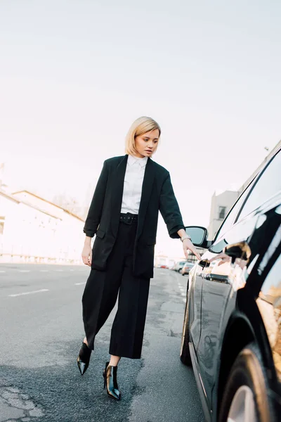 Stylish blonde woman standing near black car in parking — Stock Photo