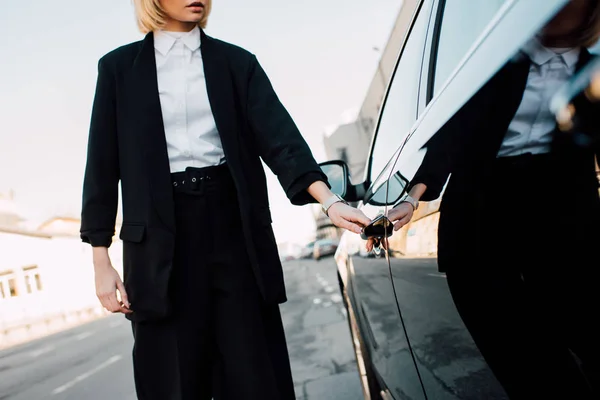 Vista ritagliata di elegante donna bionda in piedi vicino auto nera nel parcheggio — Foto stock