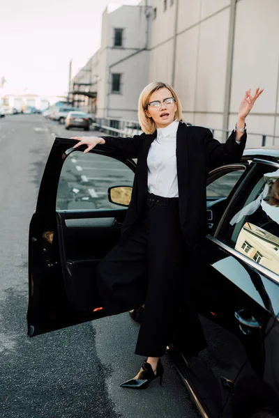 Засмучена блондинка молода жінка в окулярах стоїть біля чорного автомобіля і жестикулює в парковці — стокове фото