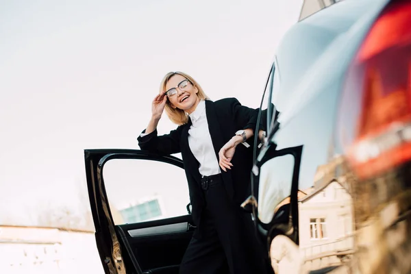 Vista de ángulo bajo de la joven rubia feliz en gafas de pie cerca de automóvil negro y gafas de tocar - foto de stock