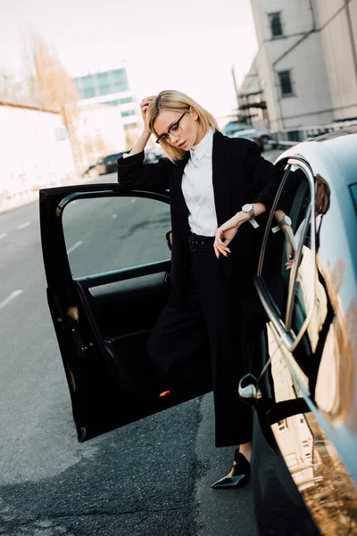 Blonde junge Frau mit Brille, die neben schwarzem Auto steht und Haare berührt — Stockfoto