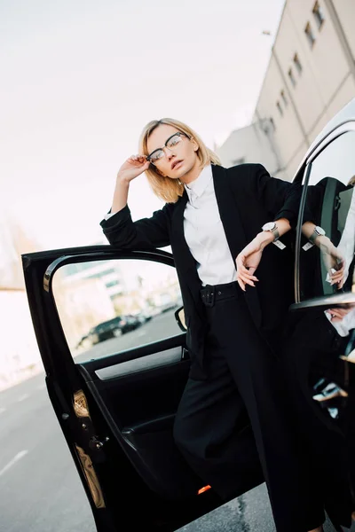Serious blonde young woman in glasses standing near black automobile and touching glasses — Stock Photo