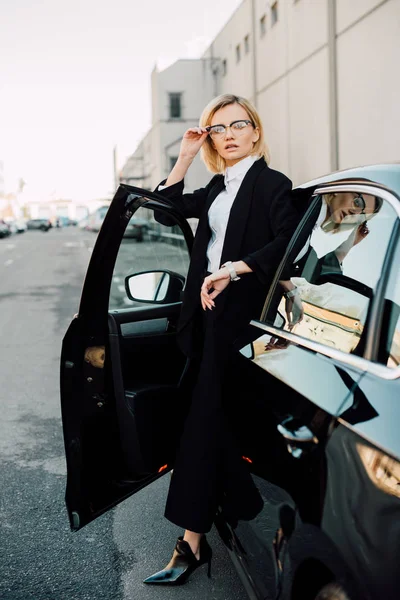 Bionda giovane donna in occhiali in piedi vicino a automobile nera e occhiali toccanti — Foto stock