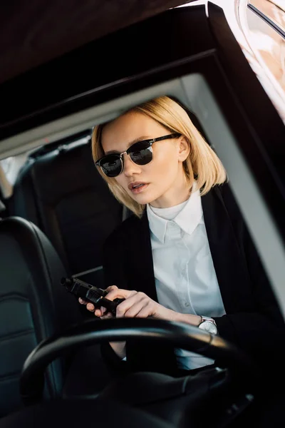 Attractive blonde girl in sunglasses holding gun in car — Stock Photo