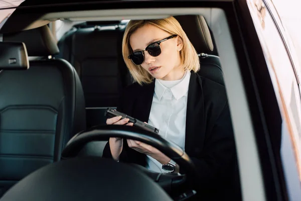 Attractive young blonde woman in sunglasses holding gun in car — Stock Photo