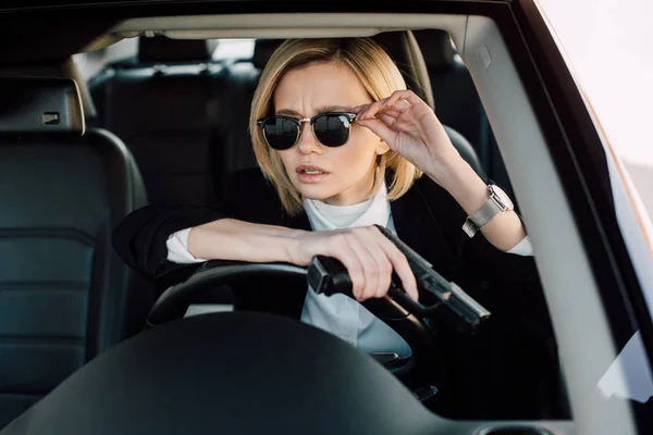 Beautiful young blonde woman holding gun at touching sunglasses in car — Stock Photo