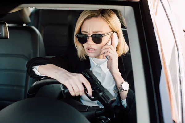 Blonde femme en lunettes de soleil parler sur smartphone et tenant pistolet dans la voiture — Photo de stock