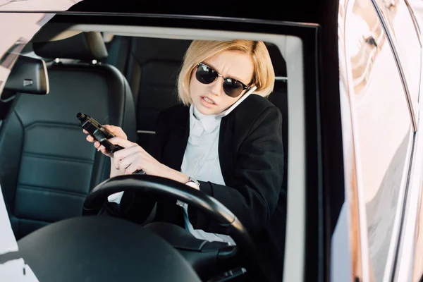 Confident young blonde woman in sunglasses talking on smartphone and holding gun in car — Stock Photo