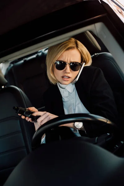 Seria joven rubia en gafas de sol hablando en el teléfono inteligente y sosteniendo arma en el coche - foto de stock