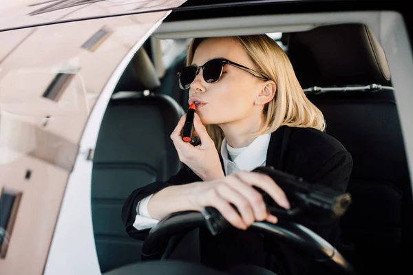 Mulher loira atraente aplicando batom enquanto segurando arma no carro — Fotografia de Stock