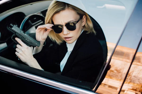 Junge blonde Frau berührt Sonnenbrille und hält Waffe im Auto — Stockfoto