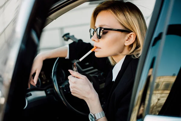 Mulher loira elegante segurando isqueiro com fogo enquanto fuma cigarro no carro — Fotografia de Stock