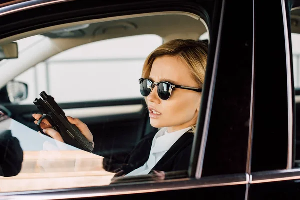Confident young blonde woman in sunglasses holding gun in car — Stock Photo