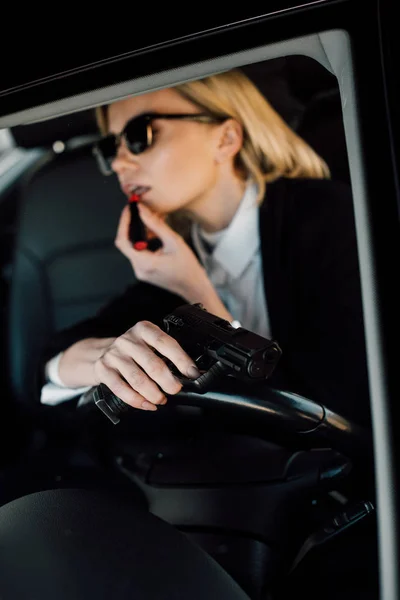 Selektiver Fokus der Waffe in der Hand einer attraktiven blonden Frau, die Lippenstift im Auto aufträgt — Stockfoto