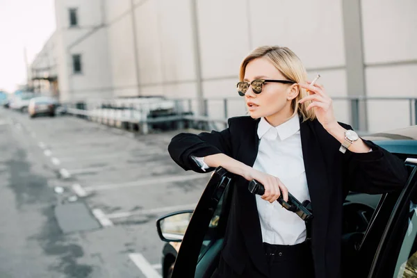 Attractive blonde woman in sunglasses holding cigarette and gun while standing near car — Stock Photo