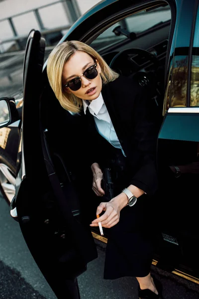 Blonde woman in sunglasses holding cigarette and gun near automobile — Stock Photo