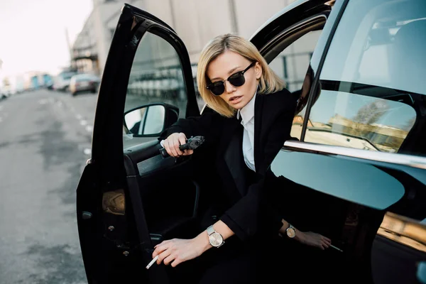 Beautiful blonde woman in sunglasses holding cigarette and gun near automobile — Stock Photo