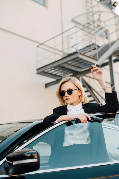 Hermosa mujer en gafas de sol sosteniendo cigarrillo y pistola cerca de automóvil - foto de stock