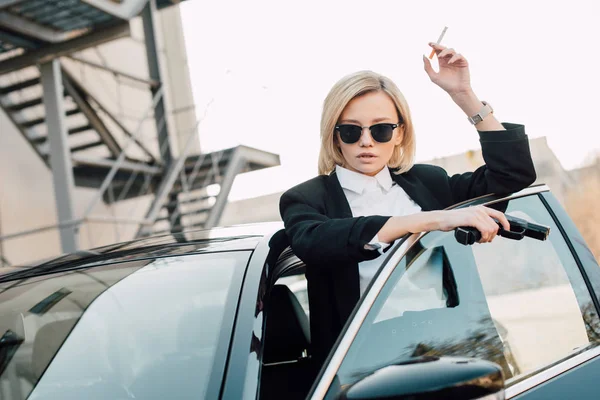 Belle blonde fumeur dans les lunettes de soleil tenant cigarette et pistolet près de l'automobile — Photo de stock